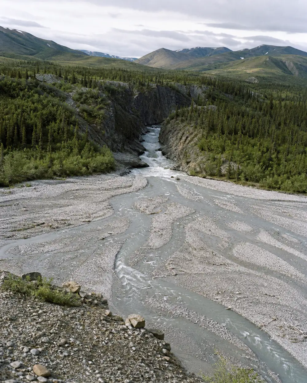 A glacial river spread across a wide gravel flat into several shallow braided channels in the foreground, with the same river squeezed into a single deep fast chute between rock banks further upstream - the same volume of water as two very different crossings | KnowOutdoor