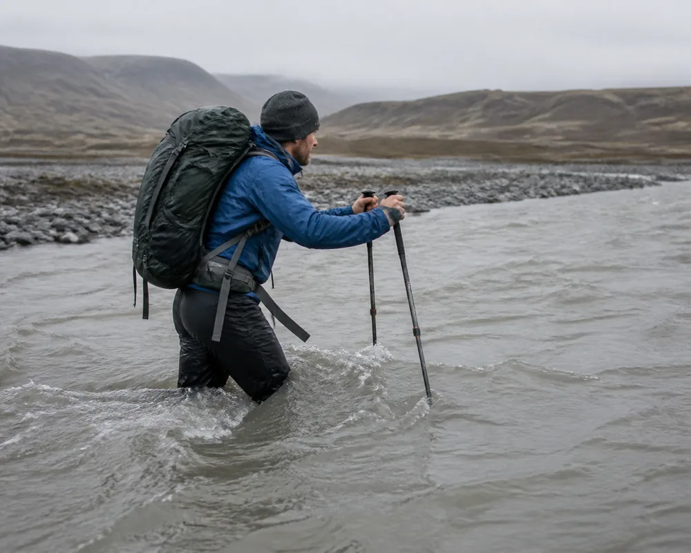 A hiker mid-river in thigh-deep grey glacial water facing upstream and angled across the flow with both trekking poles planted upstream and the hip belt hanging visibly unfastened - the two details most people get wrong | KnowOutdoor
