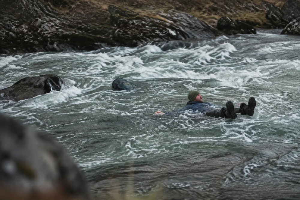 A person swept in fast whitewater lying on their back at the surface with their feet up and pointing downstream ahead of them and their chin held clear of the water, an unfastened rucksack floating loose a couple of metres behind | KnowOutdoor