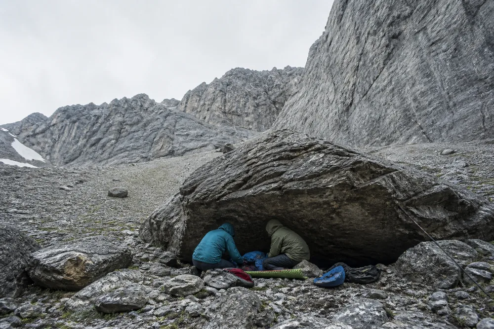 Two people tending a third lying insulated under a large overhanging boulder with the open scree slope and rock face visible beyond them - the casualty moved out of the fall line before treatment starts | KnowOutdoor