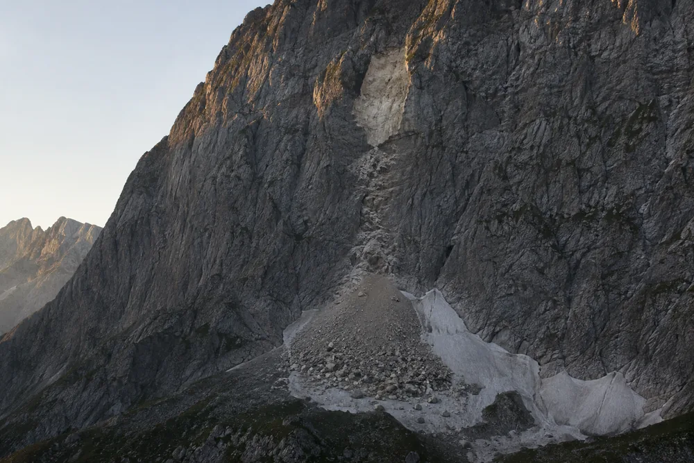 A steep alpine face in low morning light with a pale unweathered scar high on the rock and a fan of angular debris spread across the snow below it - a face recording its own recent activity | KnowOutdoor