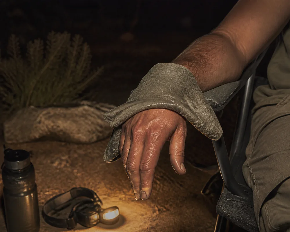 A forearm resting low on a camp chair with a damp cloth laid over the sting site, the limb clearly below chest height, lit by a warm headtorch beam at a desert campsite | KnowOutdoor