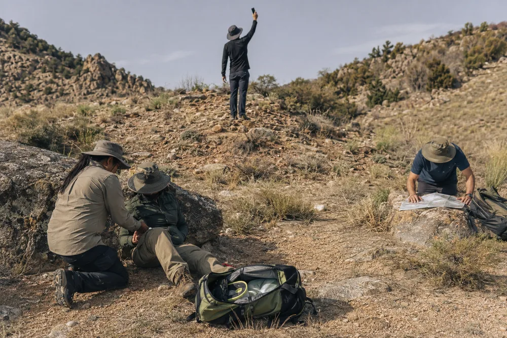 A trailside scene with one person kneeling beside a seated casualty, a second standing on a rise holding a phone up for signal, and a third crouched over a map on a rock - the four jobs running in parallel | KnowOutdoor