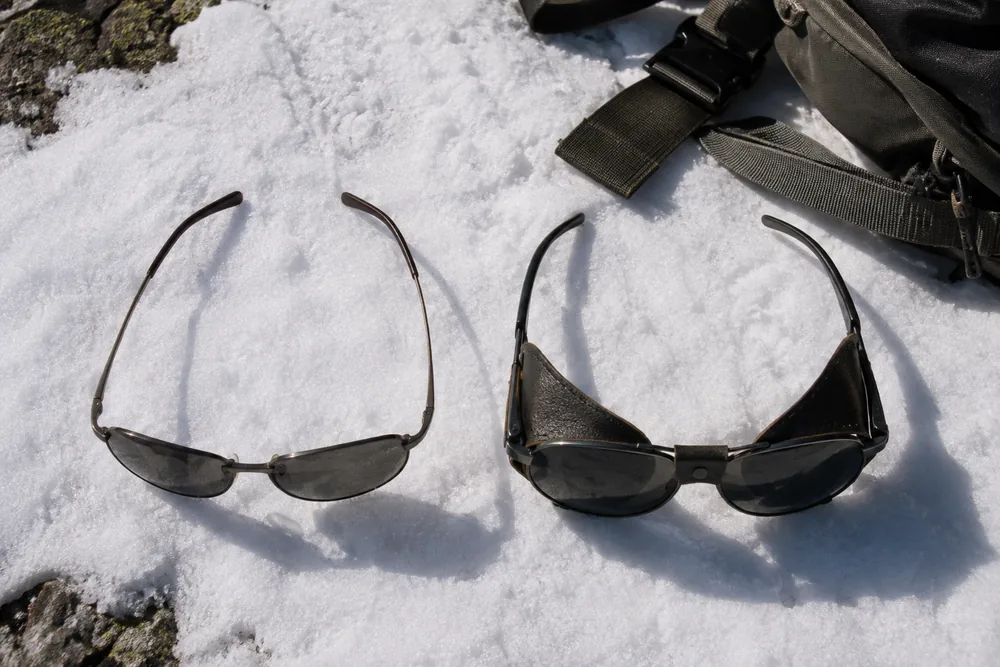 An open-framed pair of everyday sunglasses lying on snow beside a pair of wrap-around glacier glasses with solid side shields, hard sunlight showing the gap at the temple of the open pair | KnowOutdoor