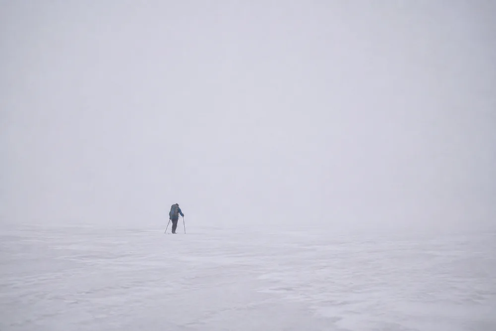 A hiker crossing a broad high snowfield under thick even cloud with no sun and no cast shadows anywhere, snow and pale sky almost the same tone - the overcast conditions that carry a full ultraviolet dose without any glare | KnowOutdoor