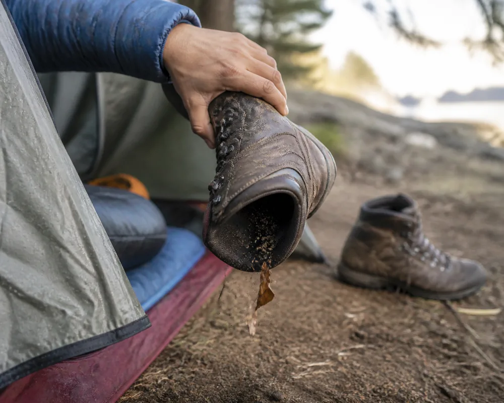A hand holding a hiking boot fully upside down and shaking it hard over bare ground outside a tent, with grit falling from the opening - shaking out rather than peering in | KnowOutdoor