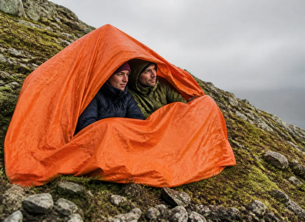 An orange group emergency shelter pulled over two seated walkers on a rocky hillside, its edges tucked under them | KnowOutdoor