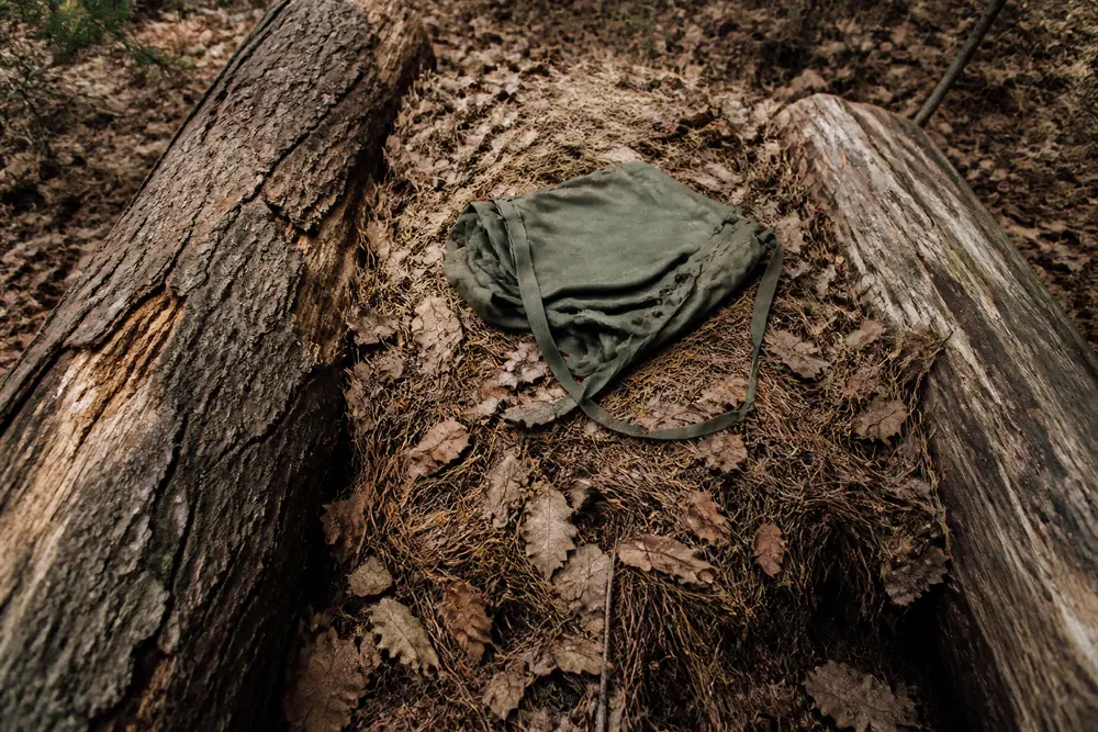 A thick heaped bed of dry leaves and conifer needles laid on woodland floor between two fallen logs, with an empty rucksack flattened on top of it | KnowOutdoor