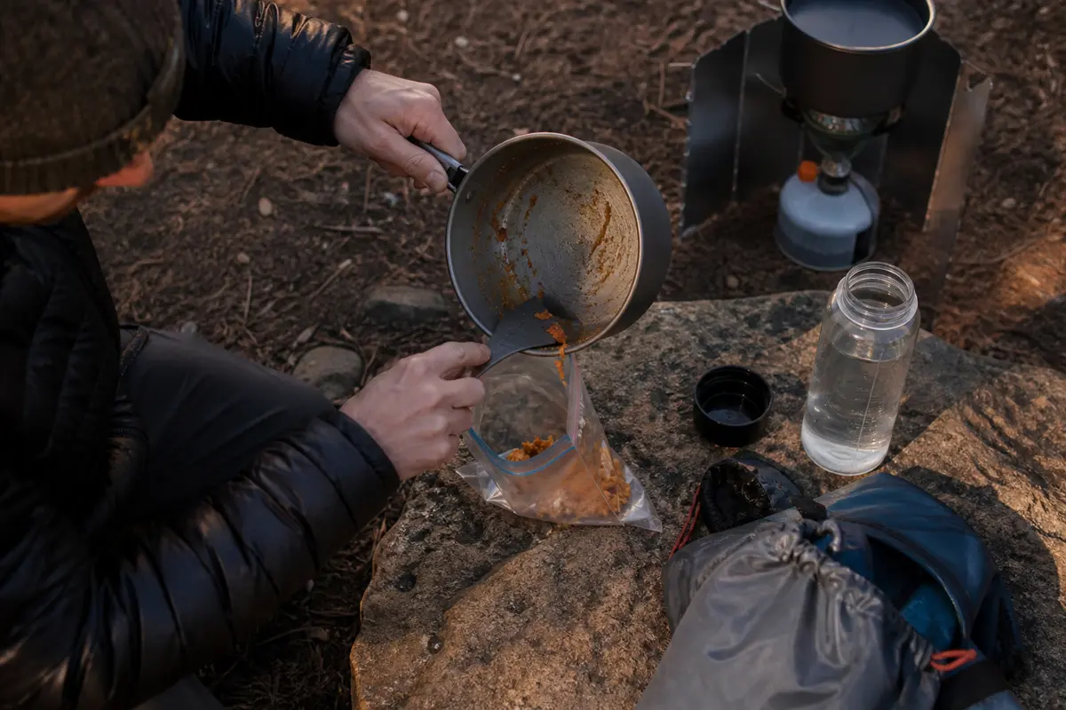 Backpacker scraping a nearly clean pot into a waste bag before using a small amount of treated rinse water away from camp
