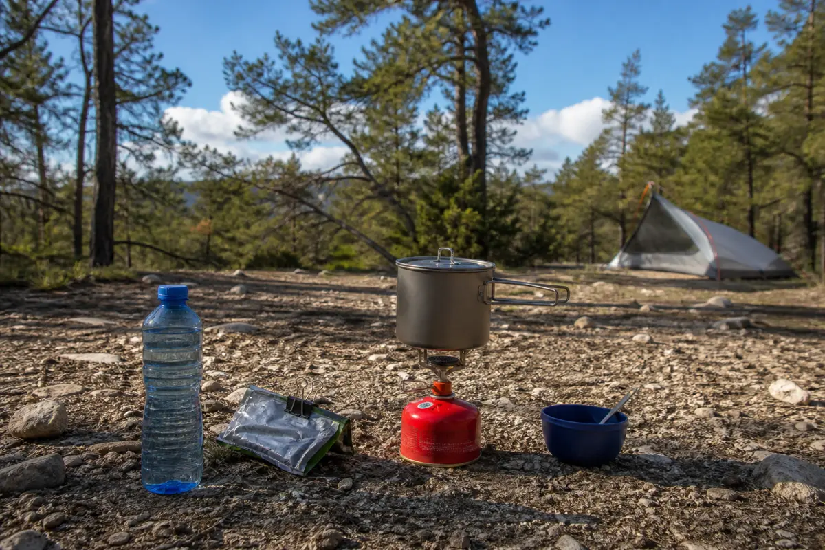 Stable backpacking stove on bare mineral ground with correctly sized pot, fuel upright, loose gear secured, and tent well outside the cooking area
