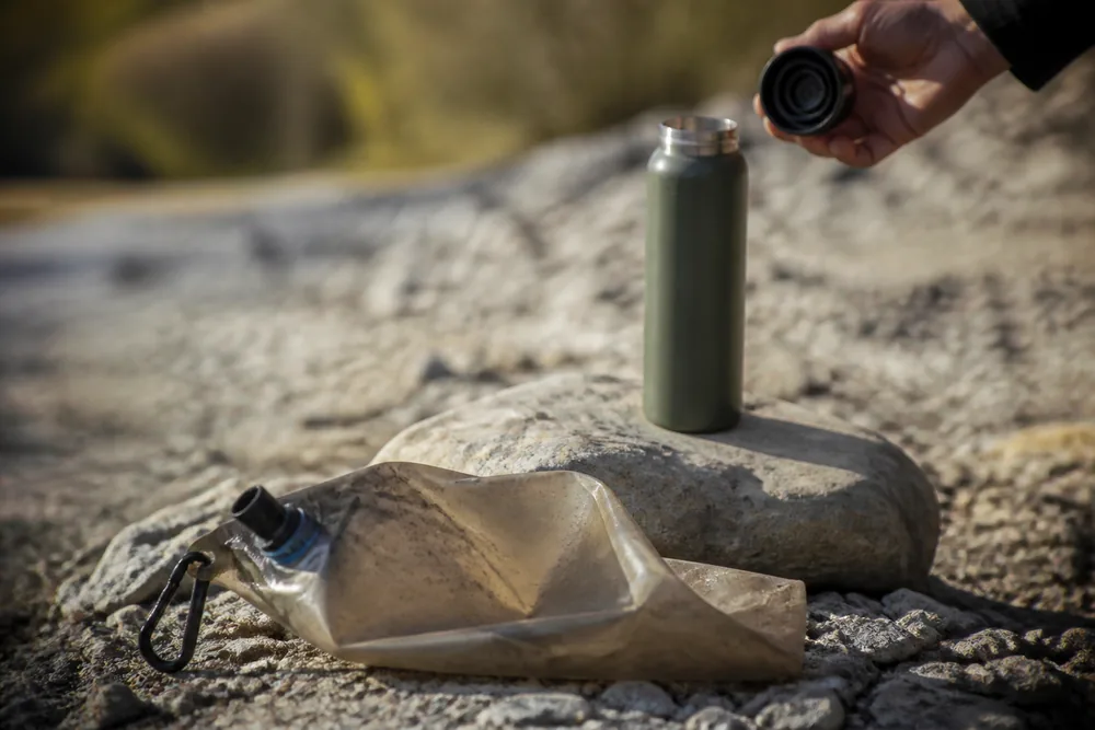 A dirty collapsible water bag separated from a clean metal bottle while a hand holds the bottle cap - Backcountry water purification | KnowOutdoor