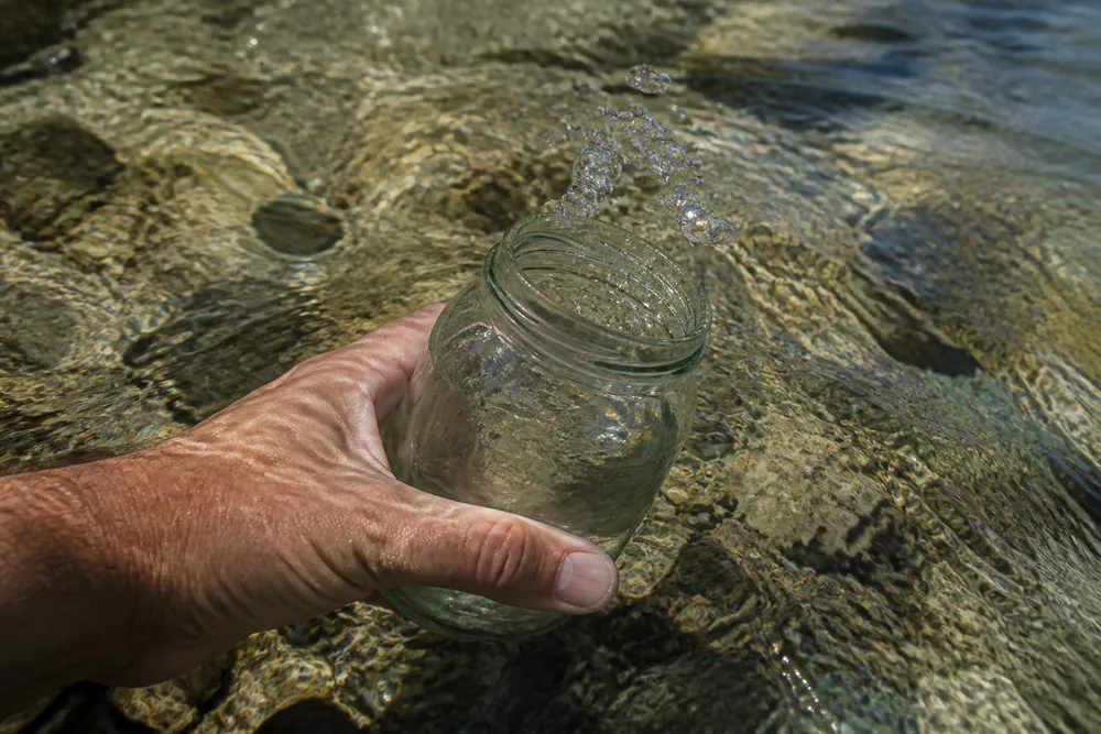 A hand holding a wide-mouth glass jar just below the surface of a clear moving stream - Backcountry water purification | KnowOutdoor