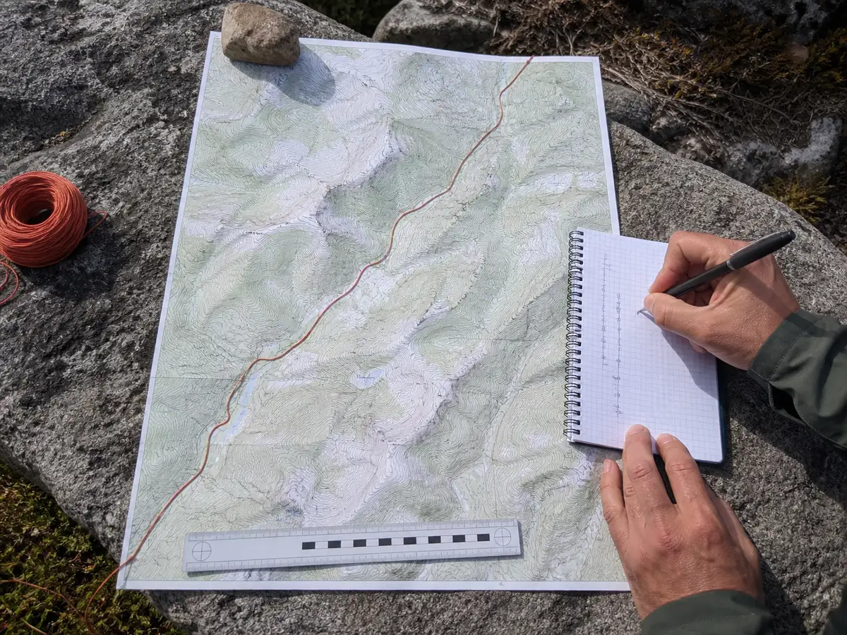 Red cord following a winding trail on a topographic map beside a metal ruler - measuring map distance | KnowOutdoor