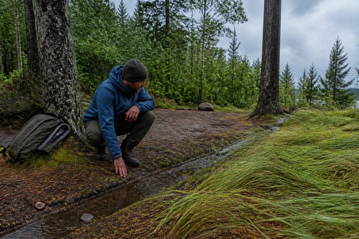 Camper checking a shallow runoff channel, wind-bent grass, sound trees, and a durable bare campsite before opening a tarp