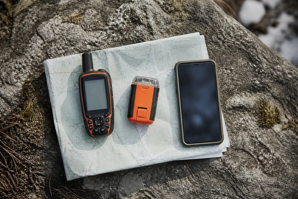 A satellite messenger, a small orange personal locator beacon and a phone laid side by side on a flat rock with a folded paper map beneath them | KnowOutdoor
