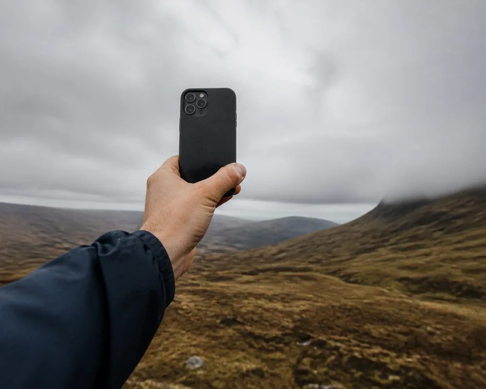 A hand holding a phone up at arm's length on an exposed ridge with empty hills behind, the screen showing no signal bars | KnowOutdoor