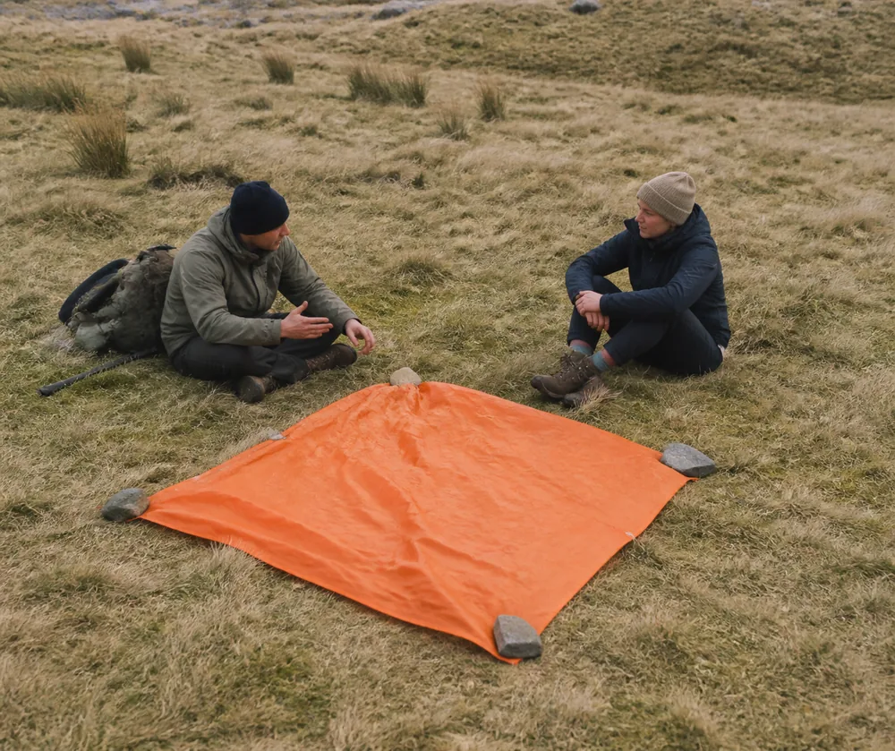 A bright orange survival bag spread flat and weighted with stones on open pale ground, with two walkers sitting beside it | KnowOutdoor