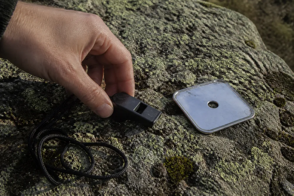 A plastic whistle on a cord and a small signal mirror lying on a lichen-covered rock, with a hand reaching for the whistle | KnowOutdoor