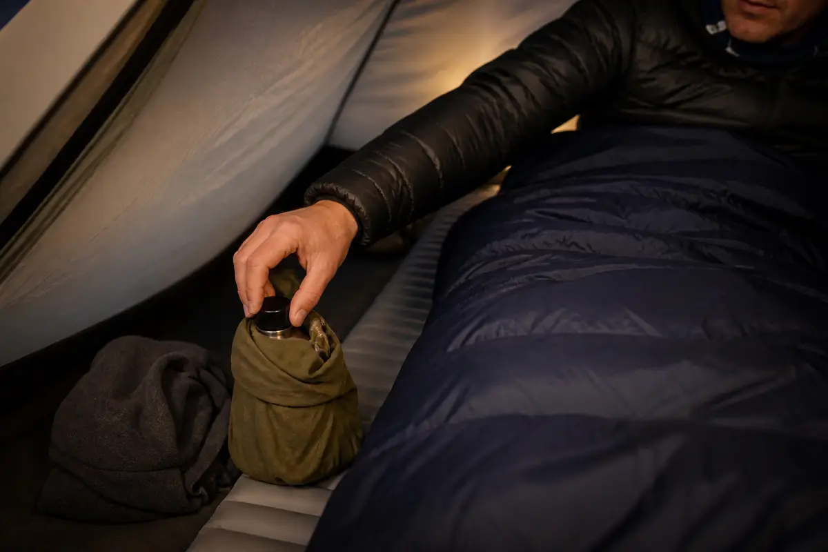 Sealed heat-safe water bottle wrapped in a dry layer near the footbox with cap checked and sleeping-bag loft left uncompressed
