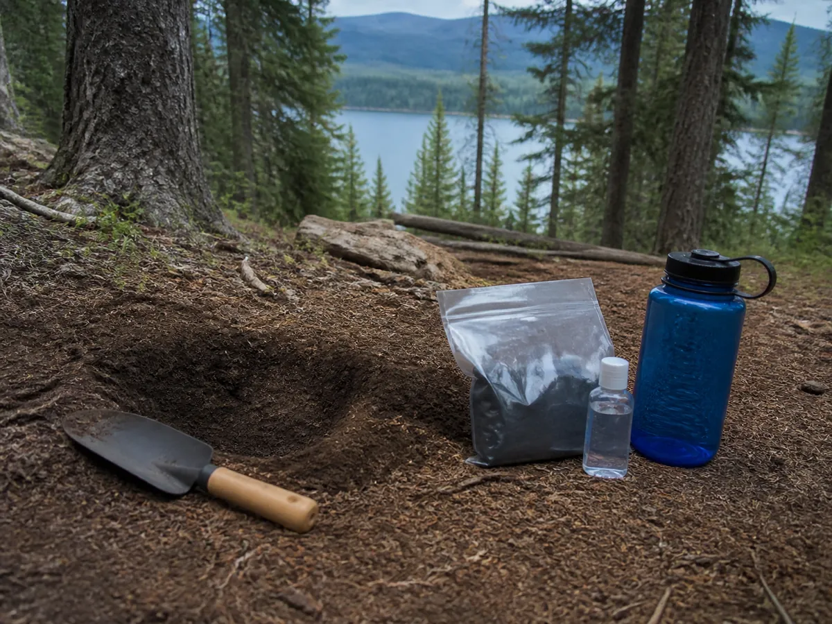 Trowel, opaque pack-out bag, sanitizer, and water bottle beside a clean cathole - outdoor waste kit | KnowOutdoor