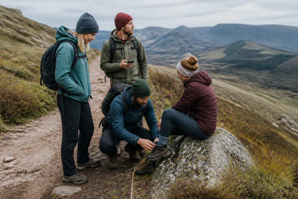 Three walkers gathered around a fourth sitting on a boulder at the side of a mountain trail, one crouching to look at her ankle and one holding a phone | KnowOutdoor