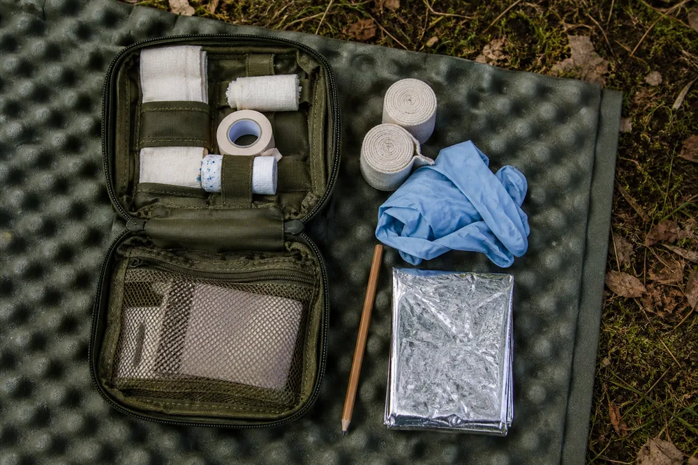 A small first aid kit opened out on a foam mat with dressings, tape, gloves, a pencil and a folded emergency blanket laid beside it | KnowOutdoor