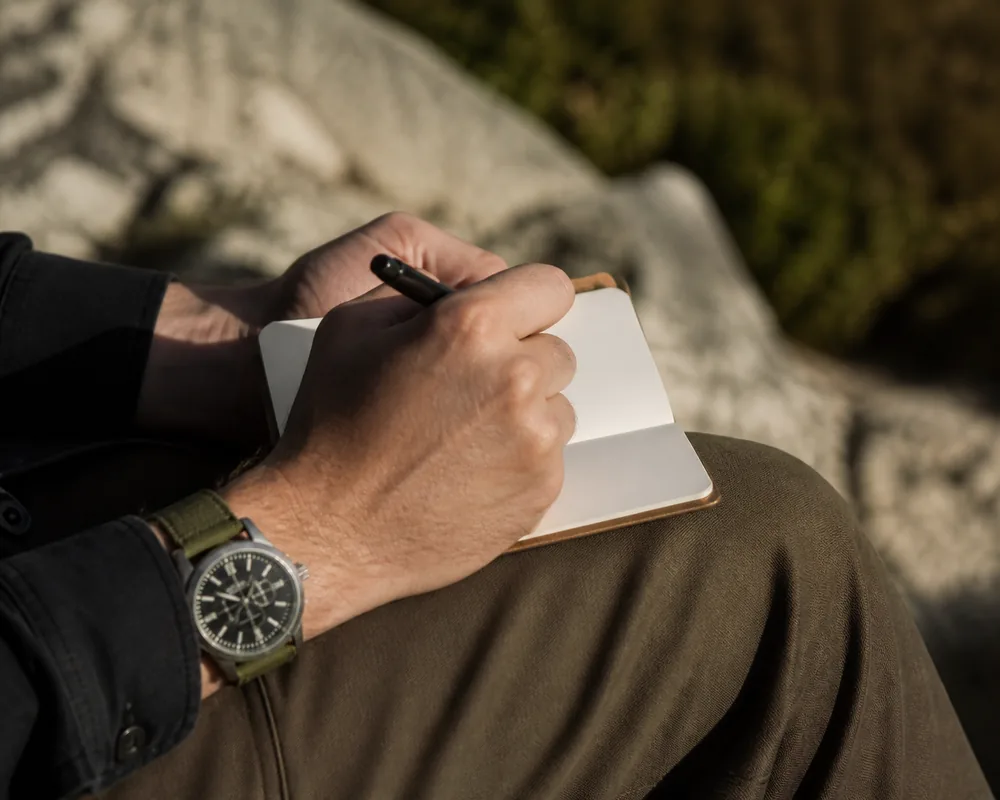A hand writing short notes in a small weatherproof notebook resting on a knee, with a wristwatch visible on the same arm | KnowOutdoor