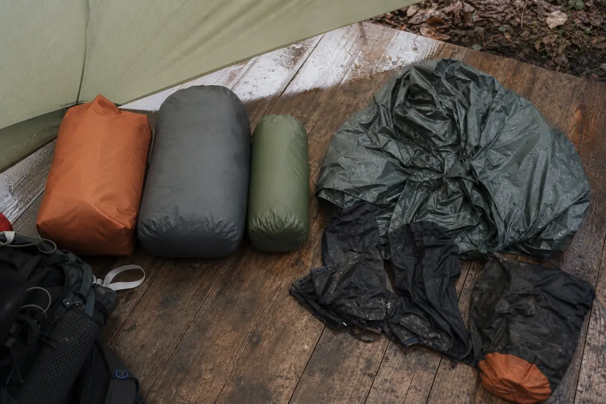 Trekking pack organized with sealed dry bags beside wet rain gear under a tarp - rainy trekking pack system | KnowOutdoor