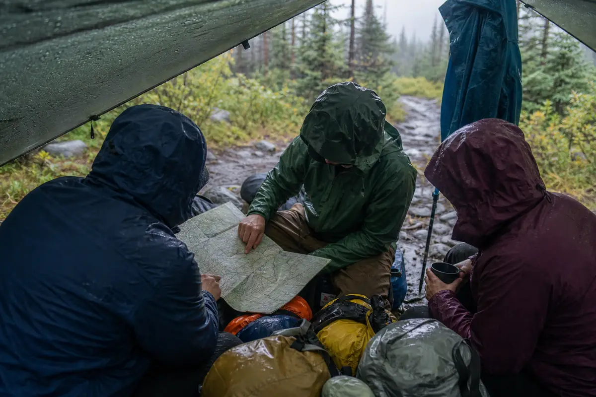 Trekkers checking damp gear, route notes, and a lower exit under morning shelter - rainy trek morning decision | KnowOutdoor
